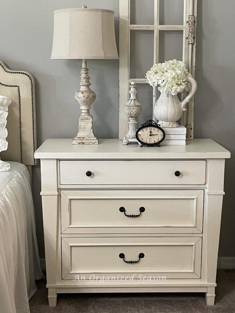 A white organized nightstand with a lamp, floral arrangement, clock, and a sculpture. 