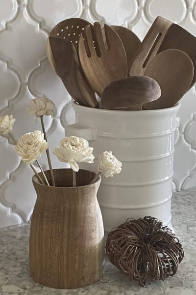 Wooden spoons in a white container, flowers in a wood vase, and a grapevine pumpkin.