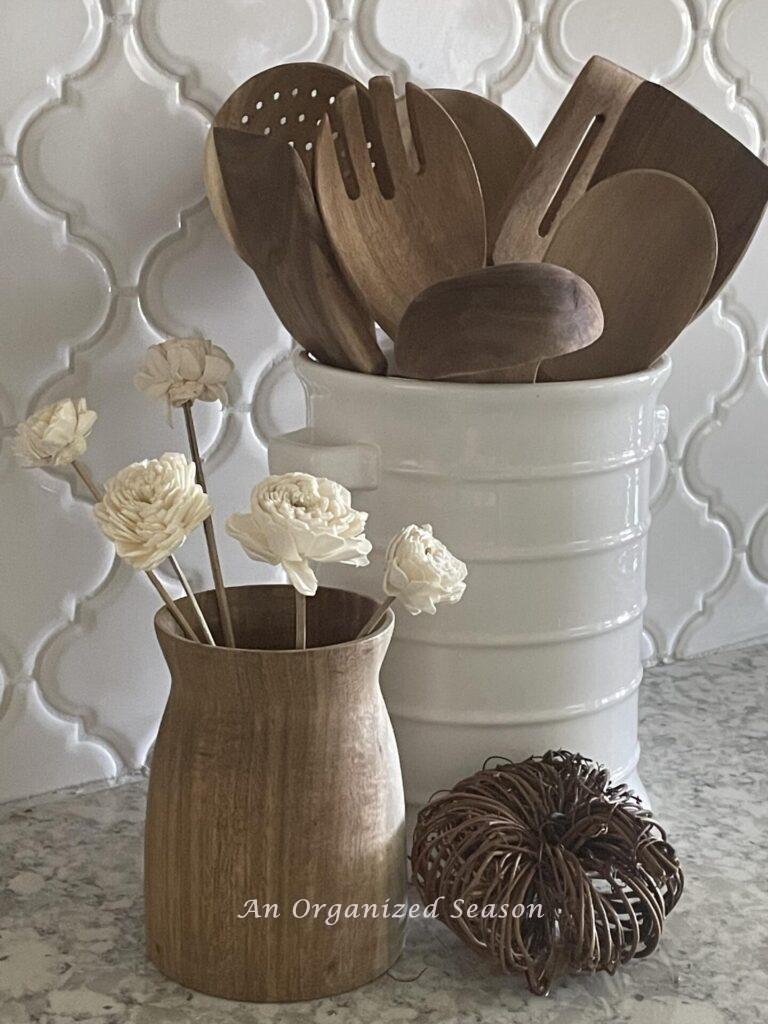 Fall kitchen decor idea #2 store wooden cooking utensils in a white container next to a wooden vase with white wood flowers and a grapevine pumpkin.