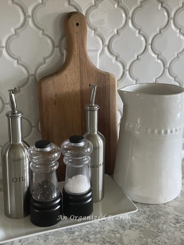 Salt, pepper, oil, and vinegar dispensers on a white tray next to a cutting board and white pitcher. 