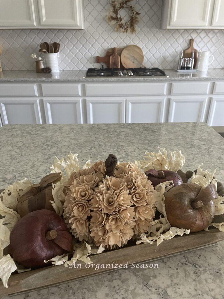 A wooden bowl holding a wood flower pumpkin and leaf-covered apples on a kitchen counter.