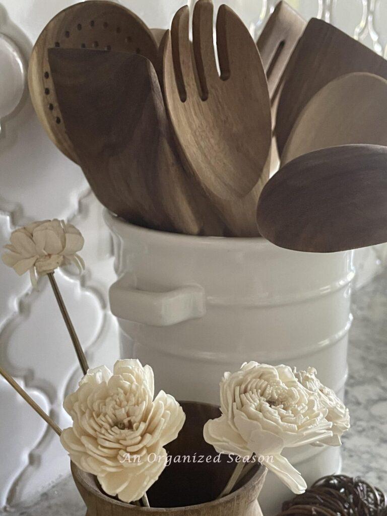 Wooden spoons in a white container next to wood vase with white wood flowers. 