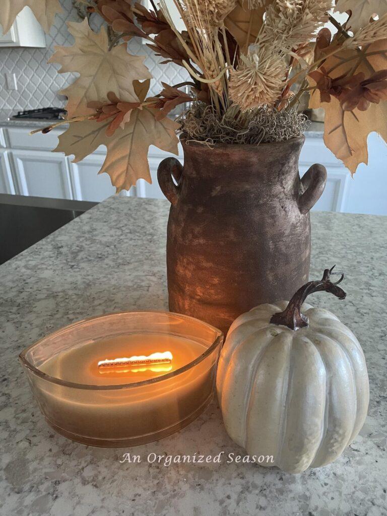 A burning candle next to a white pumpkin with a fall floral arrangement behind them. 