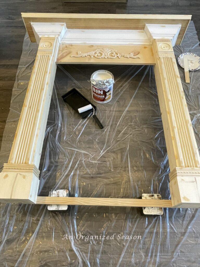 Faux fireplace mantel lying on plastic tarp near can of primer and paint roller and tray