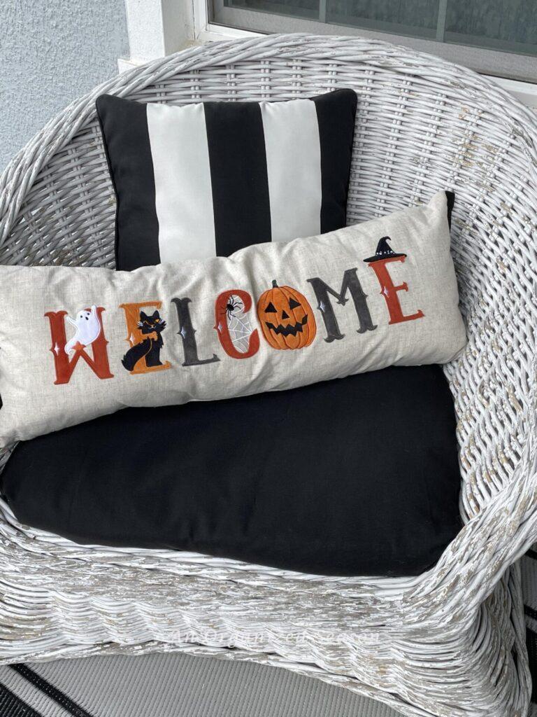 A fall "welcome" pillow sitting in a white wicker chair with a black cushion. 