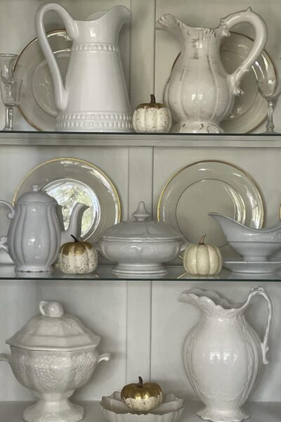 A white china cabinet filled with white dishes and pumpkins