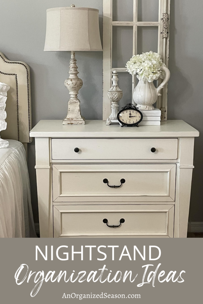 A white nightstand with a lamp, floral arrangement, clock, and sculpture.