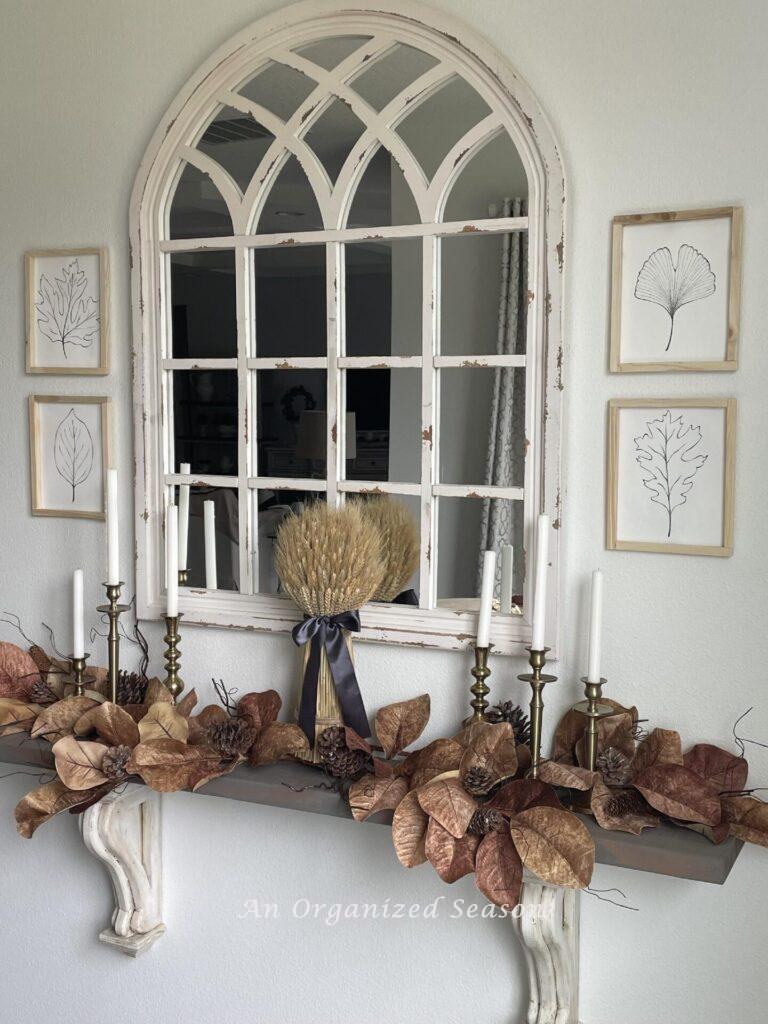 A mirror hanging above a shelf decorated with brown leaves, brass candlesticks and a wheat bundle. The mirror is flanked by two sets of leaf artwork. 