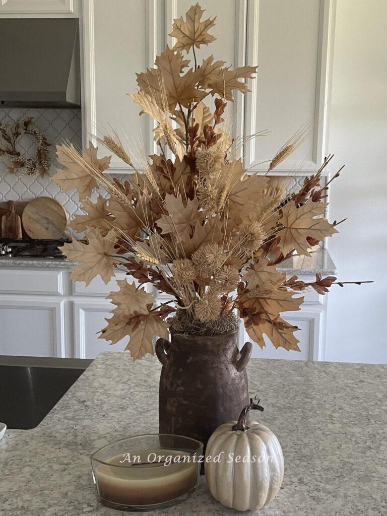 A neutral fall arrangement, candle, and pumpkin sitting on a kitchen counter. 