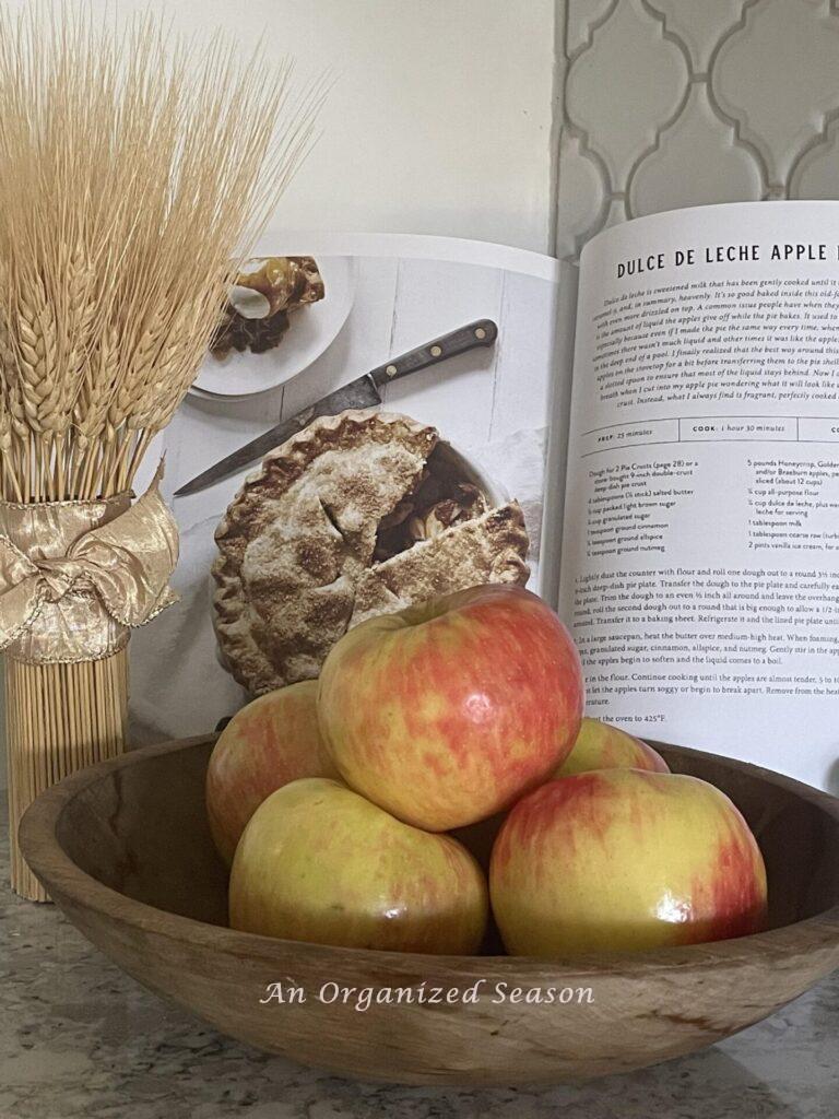 An open cookbook behind a bowl of apples and a small wheat arrangement.