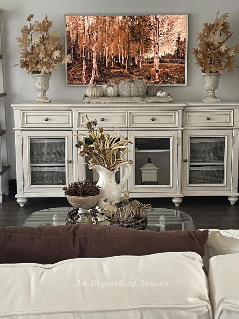 A picture frame tv displaying a fall scene is over a white console table holding a tray of pumpkins and two vases with tan leaves.