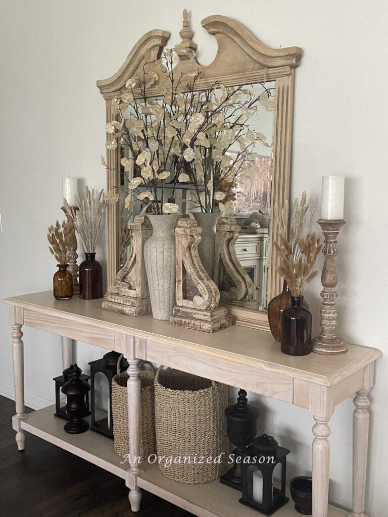 A console table holds a mirror, two candlestick holders, brown amber bottles, two corbels, three lanterns, and two baskets. 