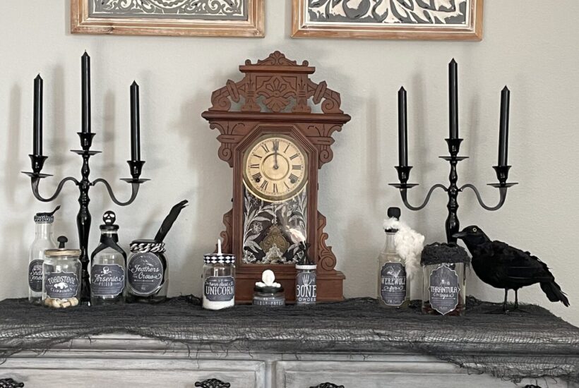 A dresser with antique clock, black candlesticks, a crow, and spooky potion bottles.