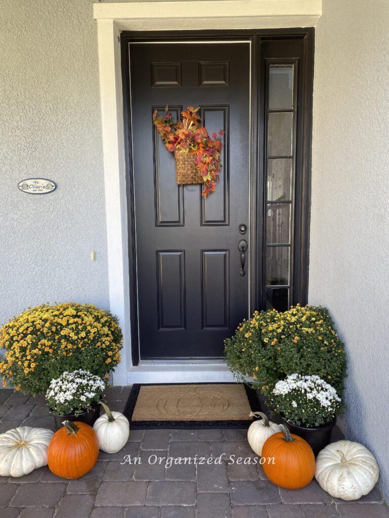 A black front door decorated with a fall basket wreath and mums and pumpkins on the porch.