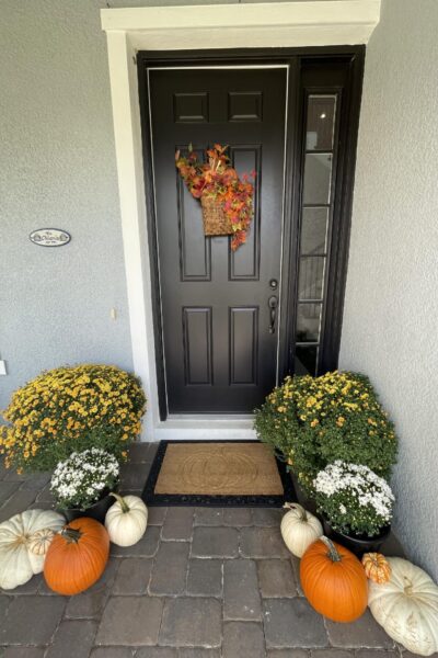 Porch decorated for fall with mums, pumpkins, a wreath, and a pumpkin doormat.