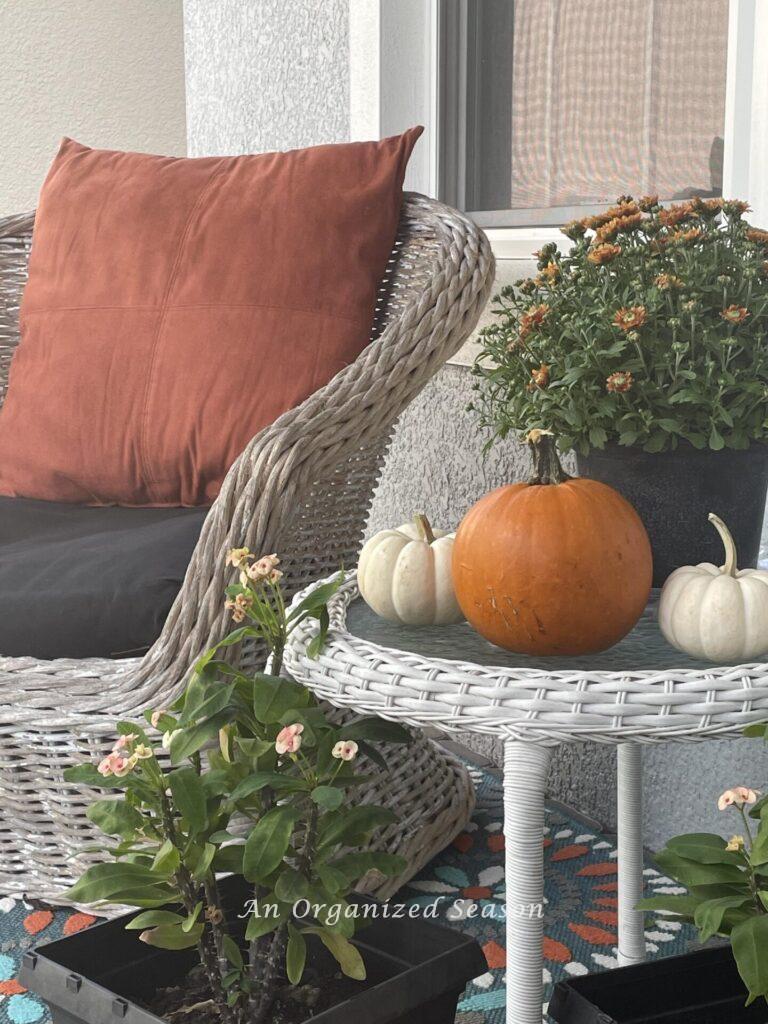 An outdoor chair with large orange pillow next to a table decorated with mums and pumpkins.