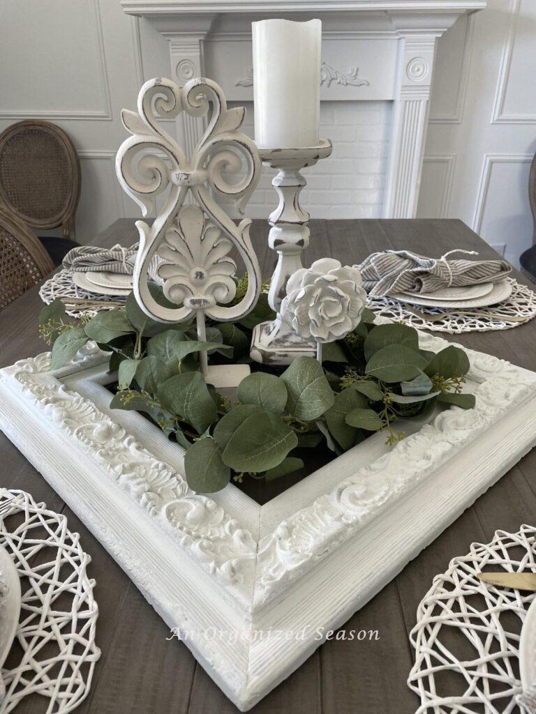 A table centerpiece with a candlestick and two white statues inside a vintage picture frame surrounded by a eucalyptus garland.