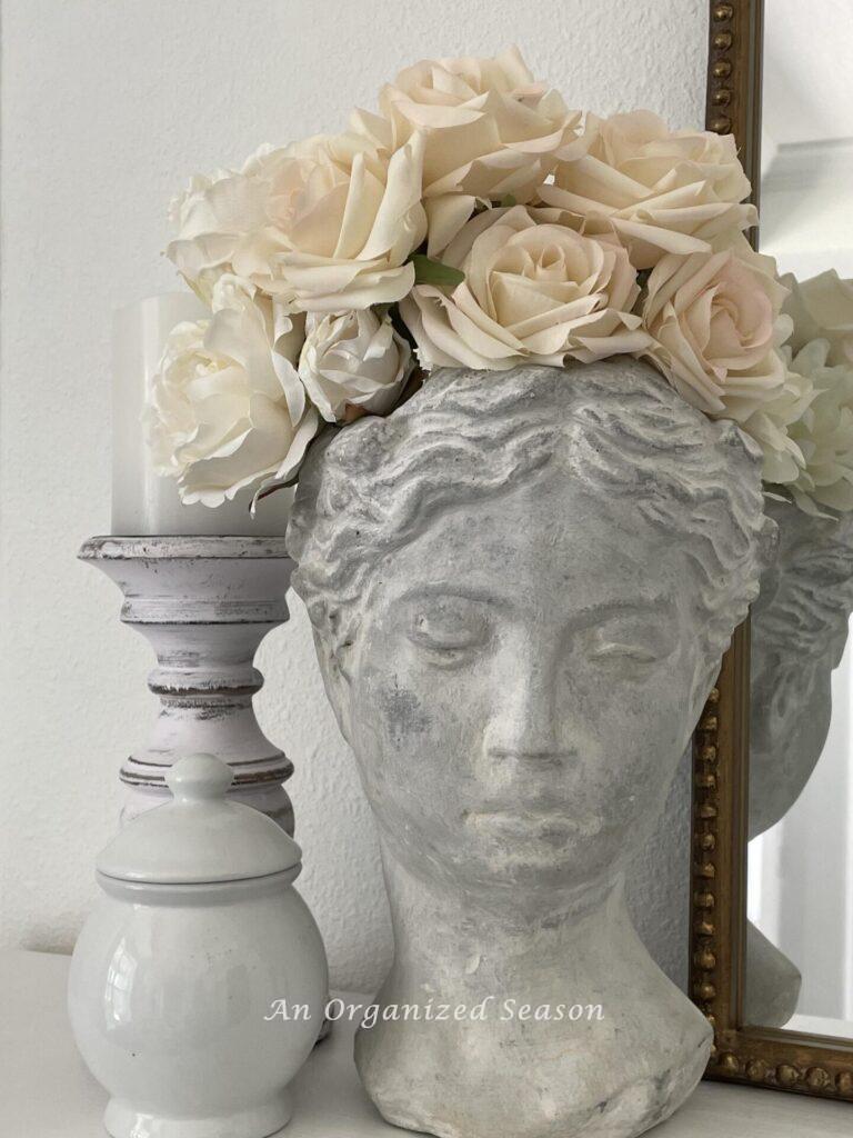 A planter bust with ivory roses, a candlestick and white dish. 