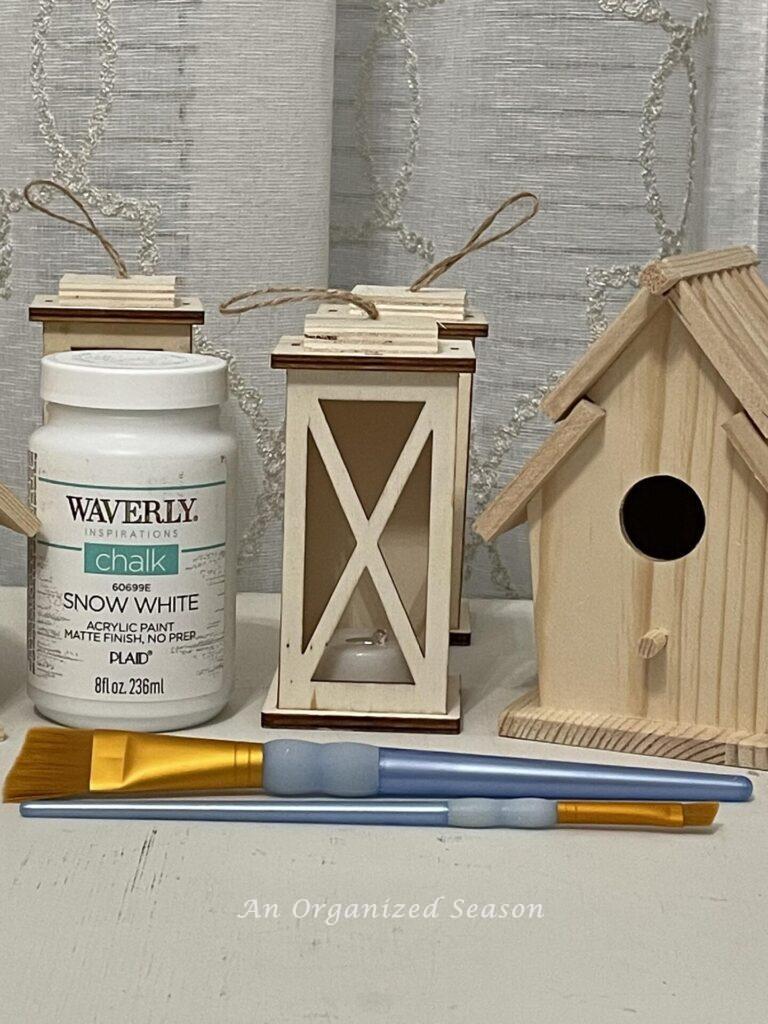 Step one to make DIY wood Christmas ornaments is to gather lanterns, birdhouses, white paint, and paintbrushes. 