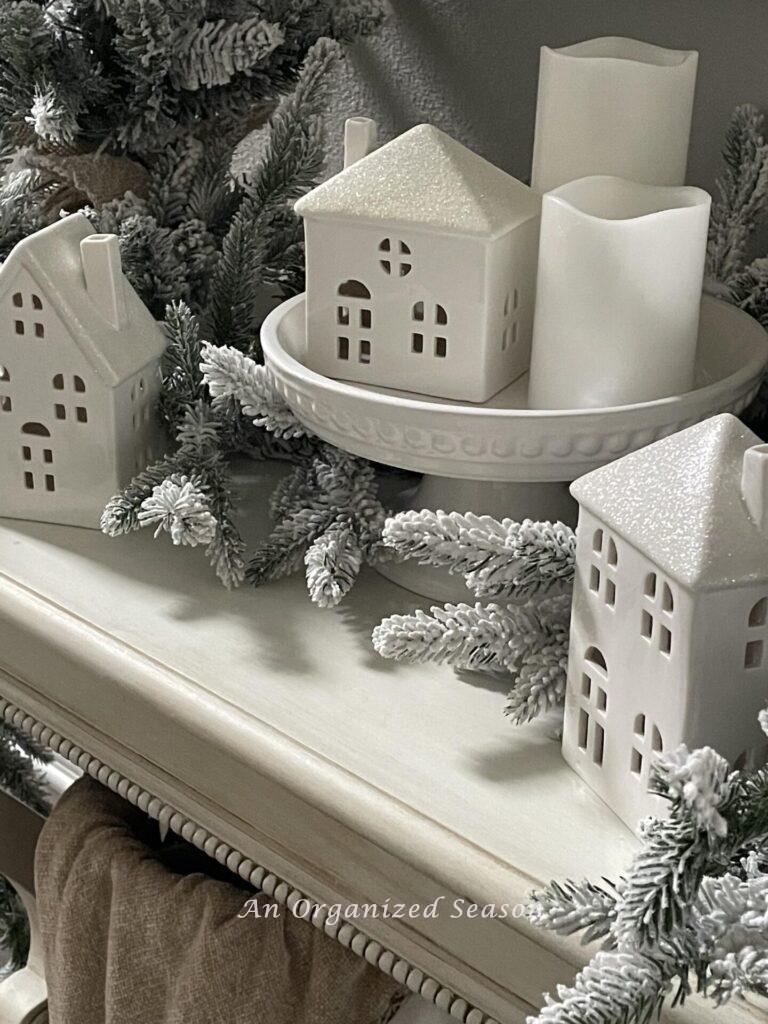 Three white ceramic houses, two pillar candles, and a white cake plate. 