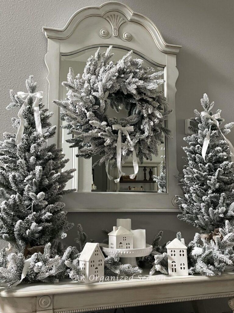 A flocked wreath, garland, and two Christmas trees surround white ceramic houses. 