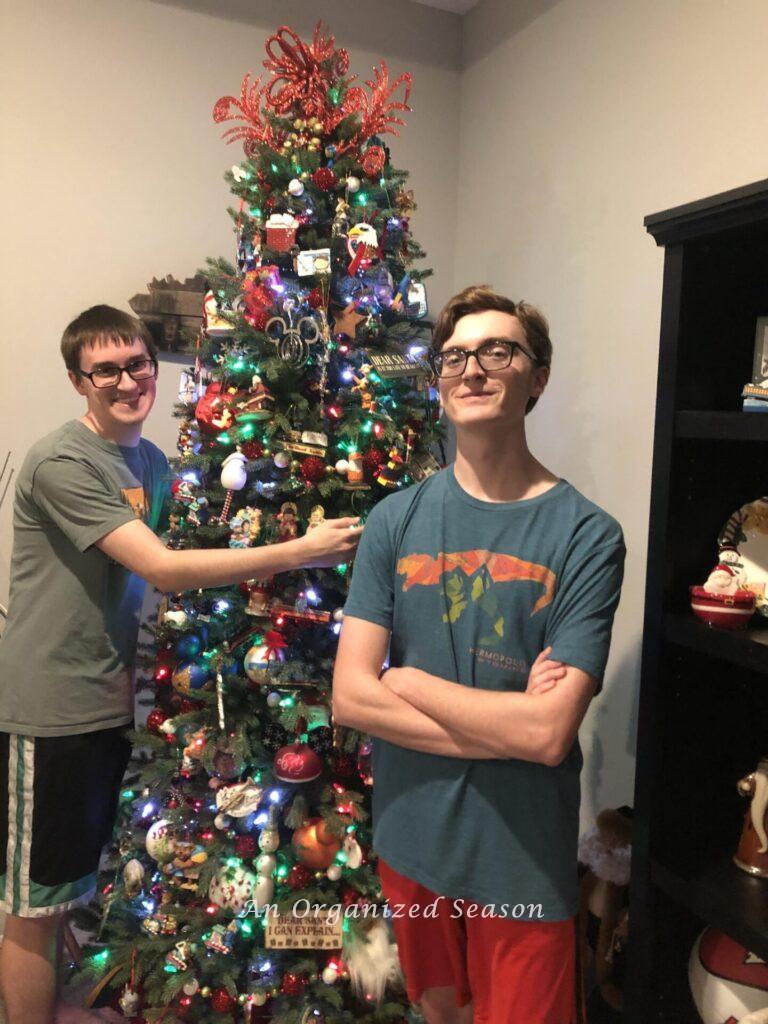 A boy hugging a Christmas tree while his brother stands in front of it.