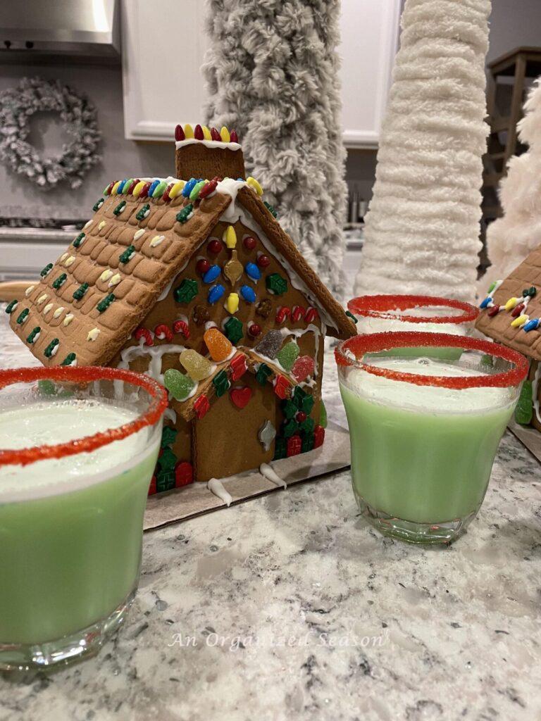 Cups of Grinch punch sitting by a gingerbread house. 