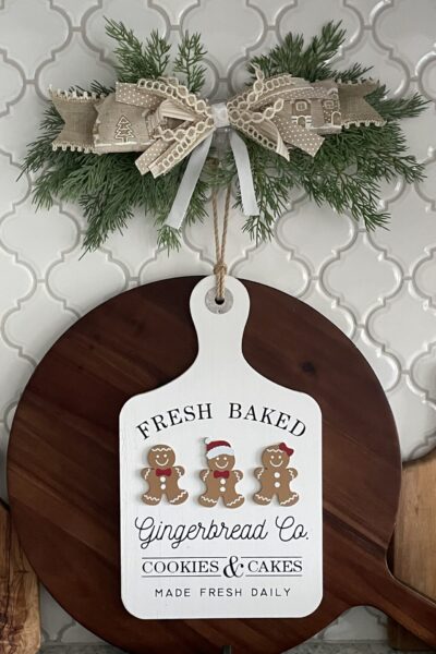 Gingerbread sign hanging over a wood cutting board with an evergreen swag and bow above it.