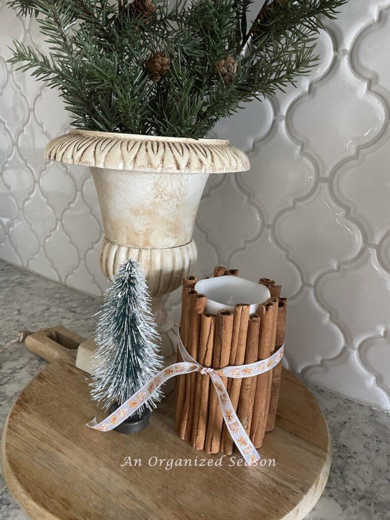 Evergreen stems in an urn sitting on a round cutting board with a bottle brush tree and a cinnamon candle. 
