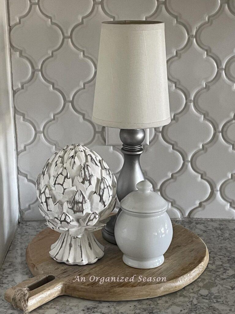 A vignette made with a cutting board, cordless lamp, artichoke statue, and white lidded bowl. 