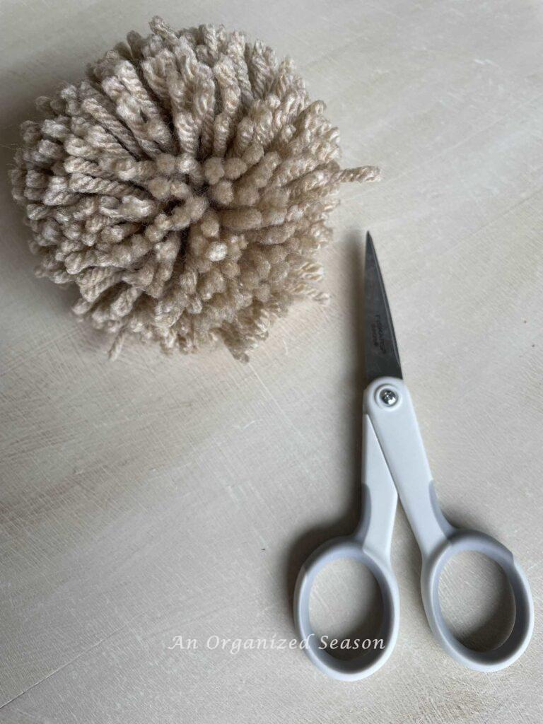 A pom pom with loose ends next to a pair of scissors. 