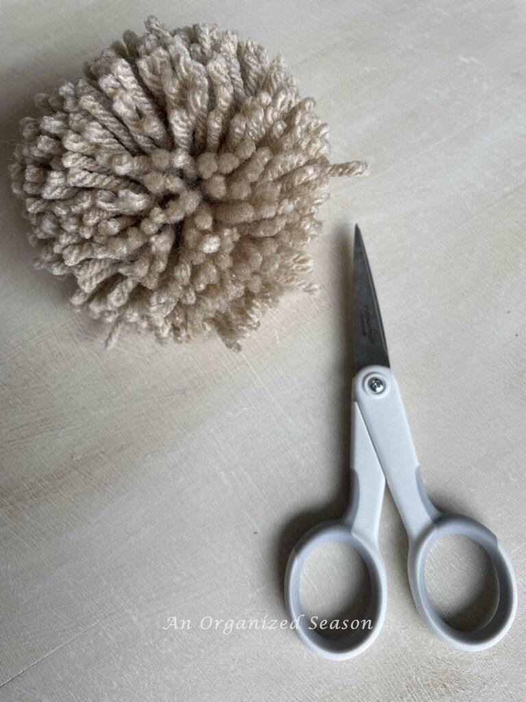 A pom pom with loose ends next to a pair of scissors.