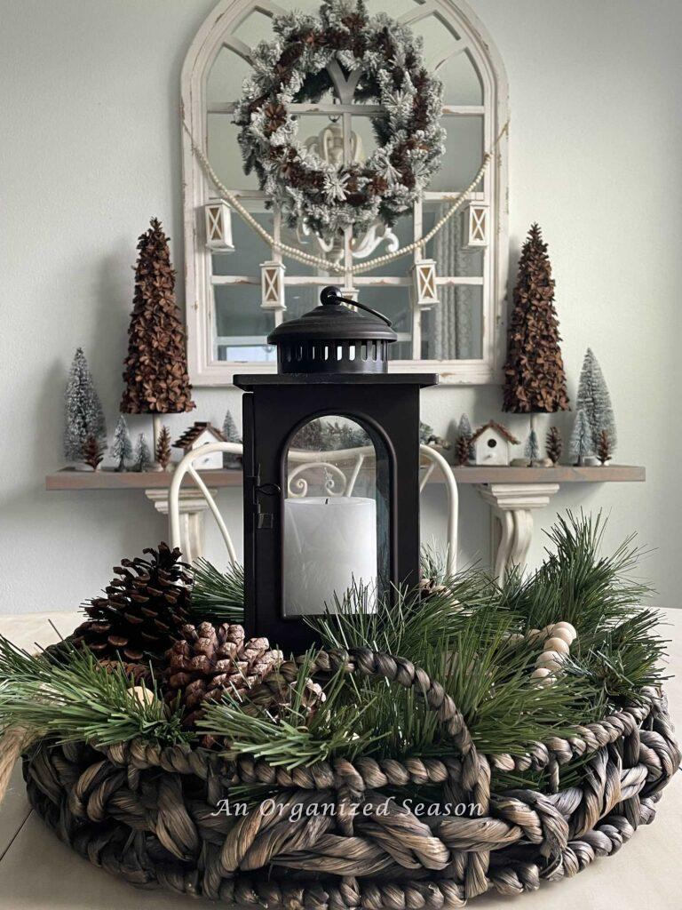 A vignette made with a round basket, evergreen pieces, pinecones, a wood bead garland, and a lantern with a candle inside it. 