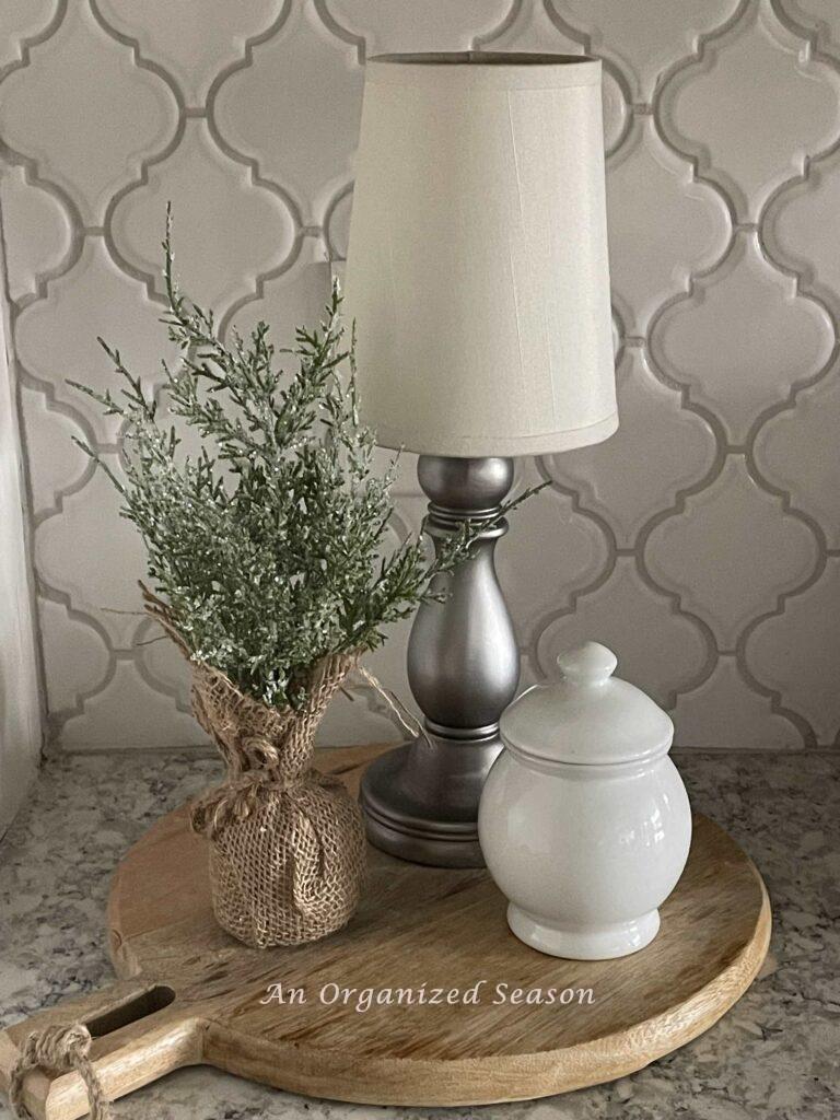 A vignette made with a cordless lamp, evergreen, and a white lidded bowl on top of a cutting board. 