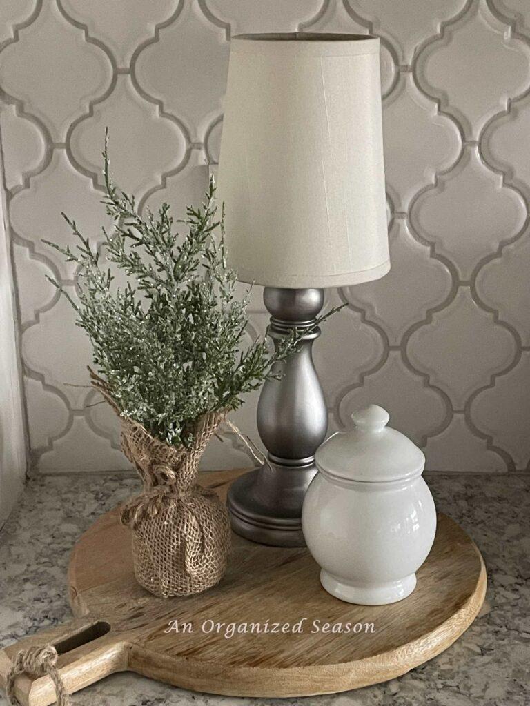 A vignette made with a cordless lamp, evergreen, and a white lidded bowl on top of a cutting board.