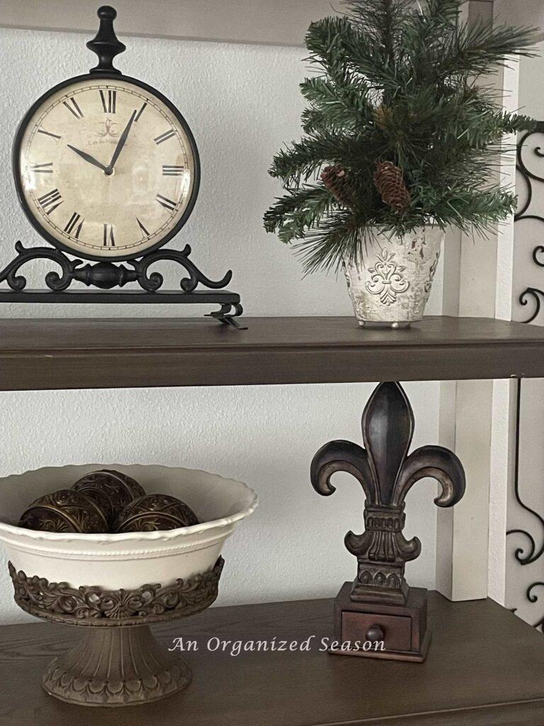Bookshelves decorated with a clock, bowl, fleur dis li, and an evergreen arrangement. 