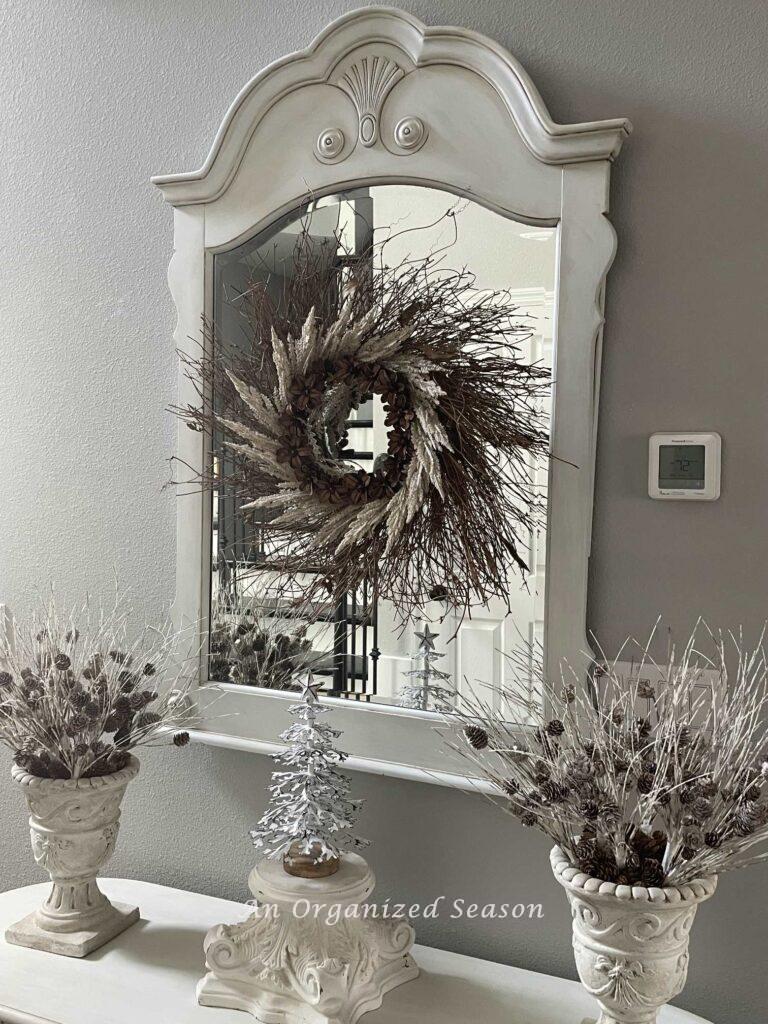A foyer table with two pinecone arrangements and a white metal tree on a pedestal with a mirror and wreath hanging above it.