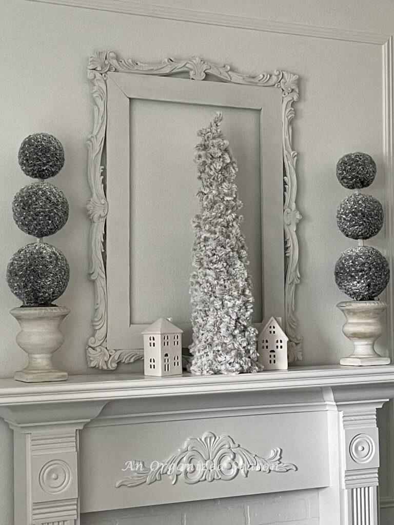 A mantle with white empty vintage frame, two topiary trees, a cone Christmas tree, and two white ceramic houses. 