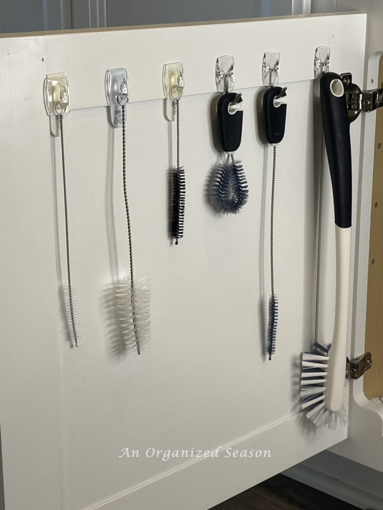 Under kitchen sink organization idea number two is to hang cleaning brushes on back of the cabinet door.