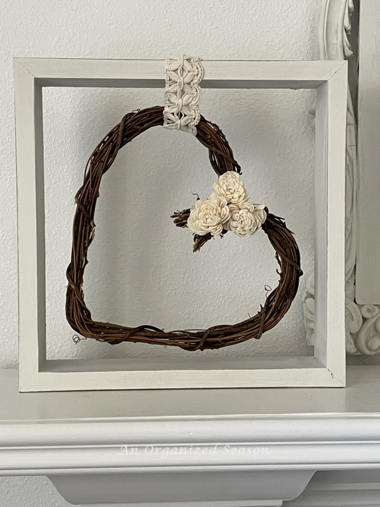 A heart grapevine wreath hanging in a square frame. 