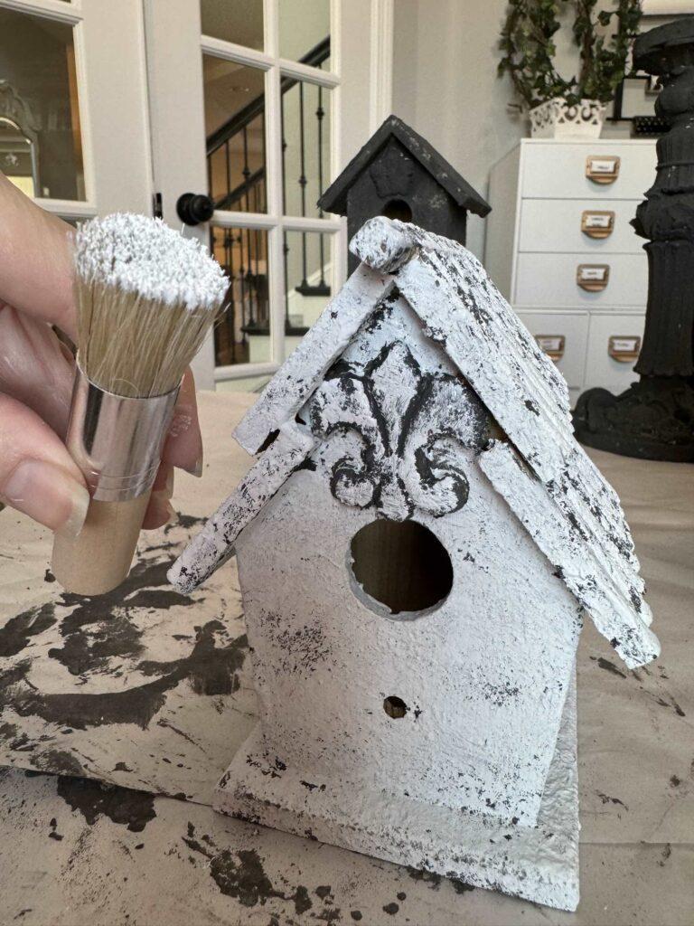 Someone holding a stencil brush tipped with white paint in front of a DIY birdhouse. 
