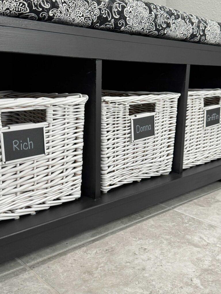 A black storage bench with three white baskets with nameplates. 