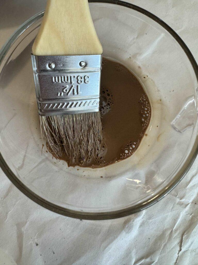 A chip brush dipped in a glass bowl with a brown paint wash. 