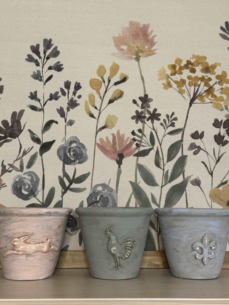 Three decorated flower pots in front of a floral canvas. 