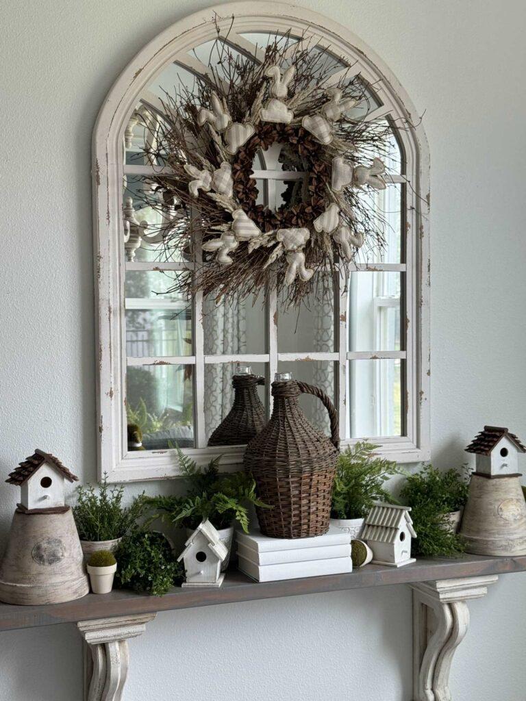 A shelf decorated with birdhouses, plants, and a large demijohn. A mirror hangs over it and it has a wreath with bunnies on it. 