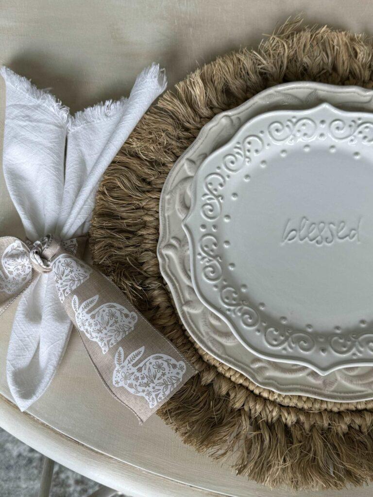 A table setting with a white salad plate that says "blessed" on a cream dinner plate and a jute placemat. A white napkin tied with bunny ribbon.