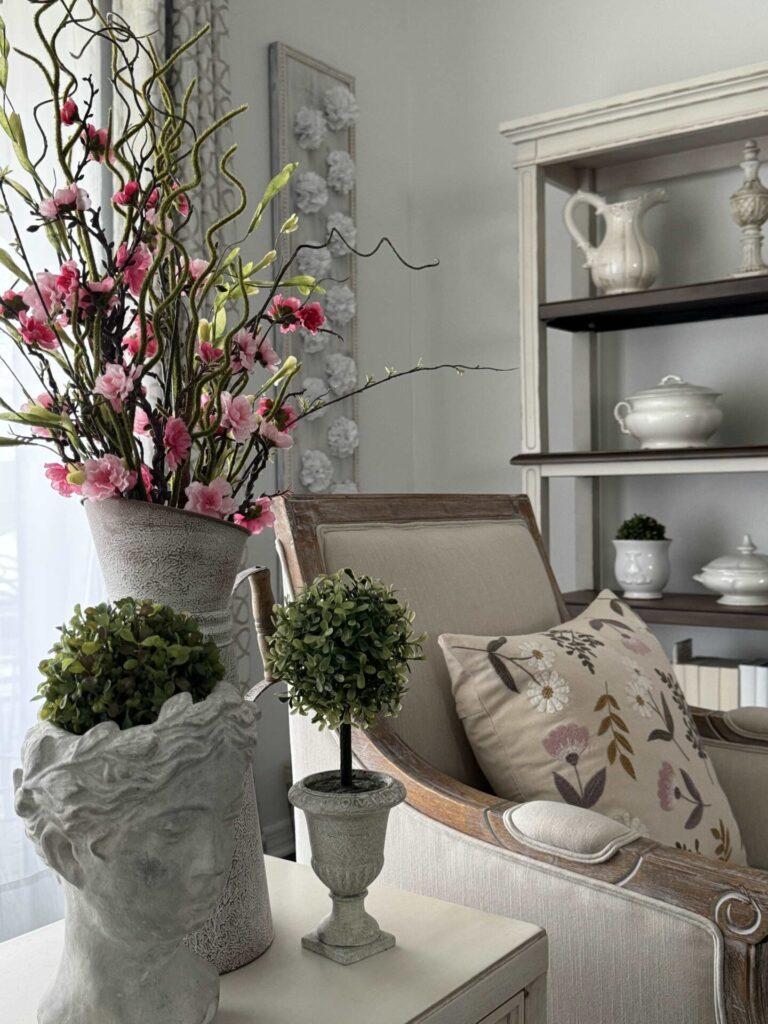 Pink flowers arranged in a metal pitcher, next to a bust statue and a boxwood topiary.