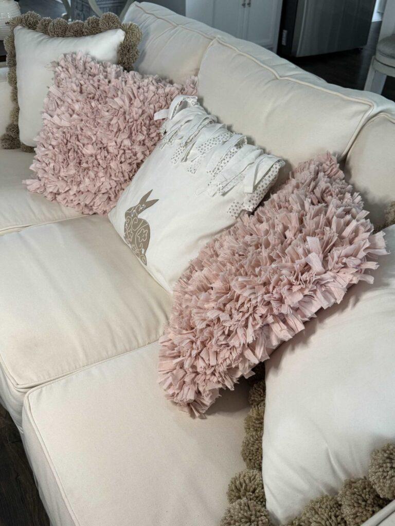 A couch lined with two pom pom pillows, two pink fluffy pillows, and a bunny pillow. 