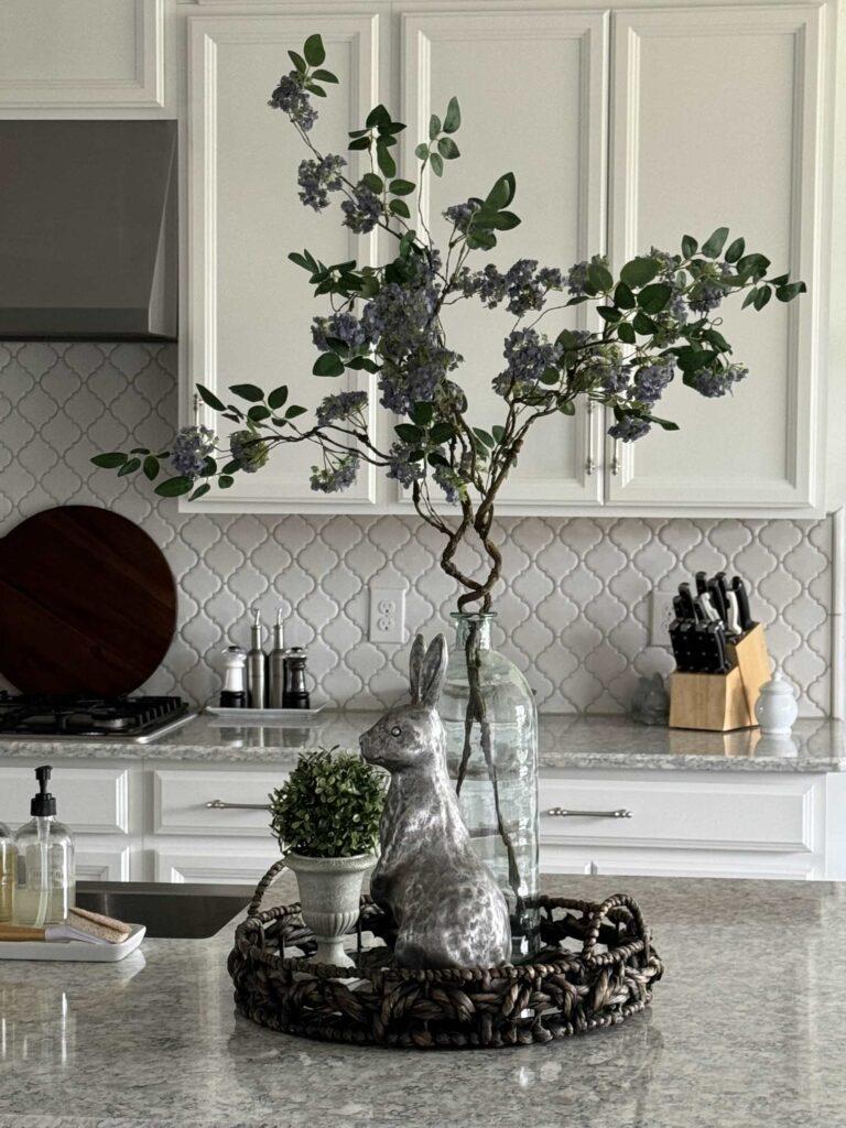 A vignette with a round basket holding a tall glass vase filled with lilac branches, a silver bunny, and a boxwood topiary.