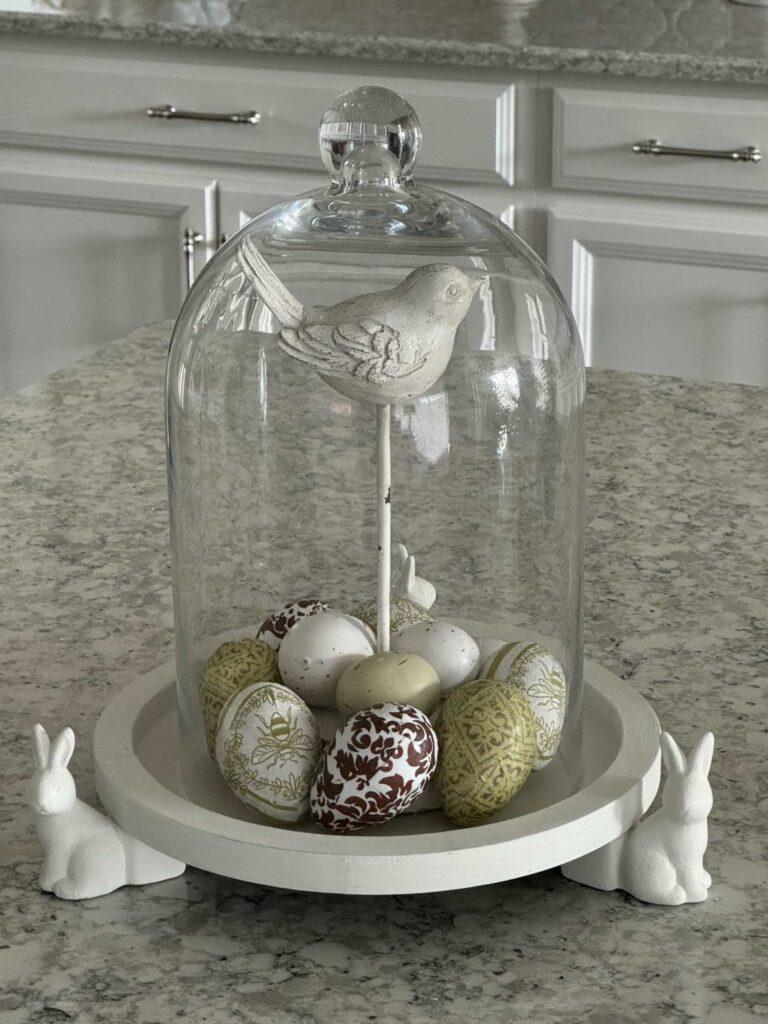 A bunny tray holding a bird statue with Easter eggs and a glass cloche covering.